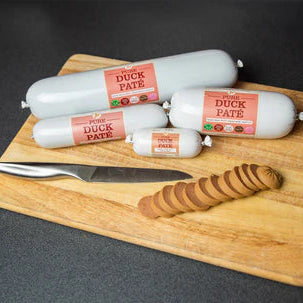 Jr Pure Duck Pâté for dogs shown in 400g, 200g, and 80g rolls on a wooden chopping board, with slices cut ready for serving.